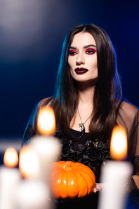 selective focus of woman with black makeup holding pumpkin near burning candles on blue, halloween concept