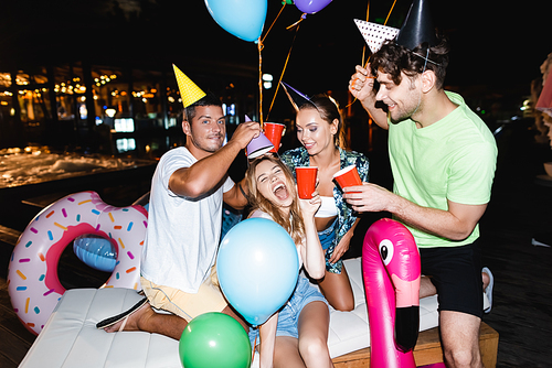 Friends in party caps holding disposable cups near swimming pool at night