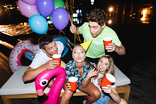 Young woman in party cap  near friends with disposable cups and balloons at night