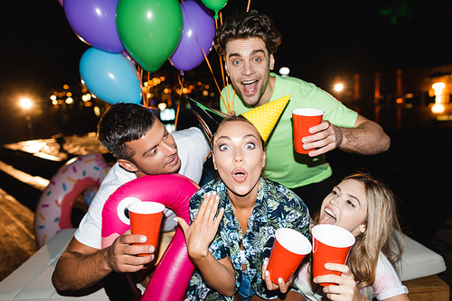 Excited woman holding disposable cup near friends with balloons at night