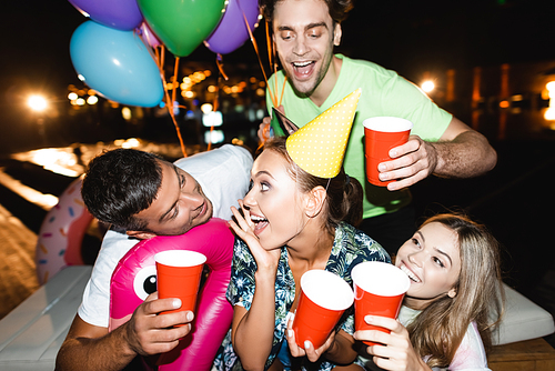 Selective focus of young woman in party cap holding disposable cup near friends with balloons at night