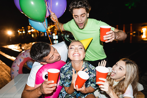 Selective focus of excited friends holding disposable cups and balloons near woman in party caps at night