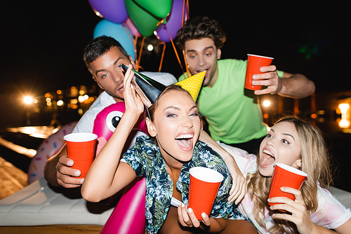 Selective focus of excited woman wearing party caps near friends with disposable cups and balloons outdoors at night