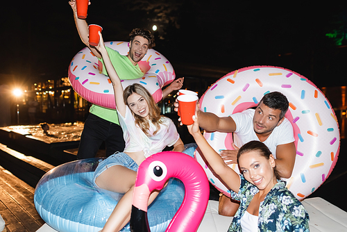 Young friends with disposable cups and swim rings  near pool at night