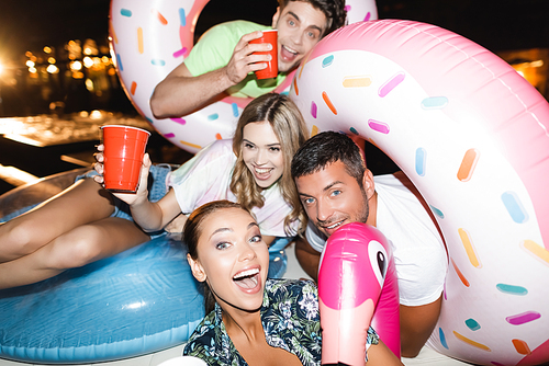 Selective focus of excited friends with swim rings holding disposable cups during party at night