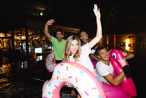 Selective focus of young woman holding swim ring and waving hand near friends during party at swimming pool at night