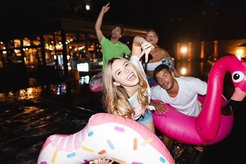 Selective focus of young woman showing peace near friends with swim rings during party at night