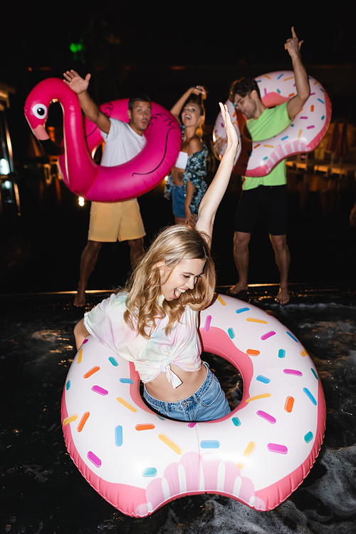 Selective focus of excited woman with swim ring standing in pool near friends at night