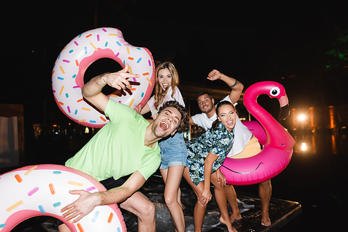 Selective focus of excited young friends with swim rings standing near pool at night