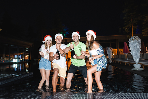 Friends in santa hats holding glasses and champagne bottles in swimming pool at night