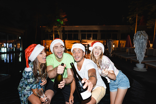 Selective focus of excited friends in santa hats holding bottles of champagne at night outdoors