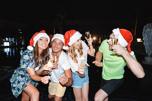 Friends in santa hats holding sparklers and champagne near swimming pool at night