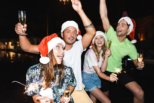 Selective focus of friends in santa hats holding glasses of champagne and sparklers at night