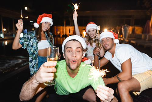 Selective focus of man in santa hat holding sparkler and glass of champagne near friends during party at night