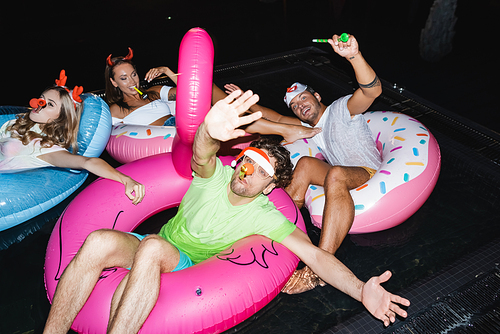 Selective focus of man with party horn waving at camera near friends in swimming pool at night