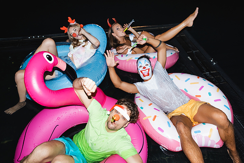 Friends blowing in party horns on swim rings in swimming pool at night