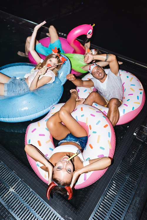 High angle view of woman in party headband with blow tickler swimming on ring near friends in pool at night