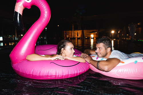 Selective focus of young couple looking at each other on swim rings in pool at night