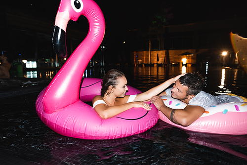 Young woman touching boyfriend while swimming in ring in pool at night