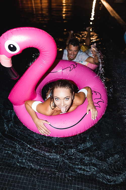 Overhead view of woman sticking out tongue near boyfriend in swimming pool at night