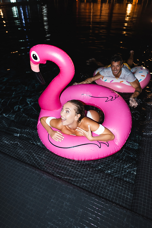 Selective focus of excited woman in swim ring looking away near boyfriend in pool at night