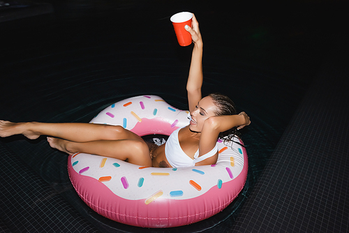 Brunette woman holding disposable cup while swimming in ring in pool at night