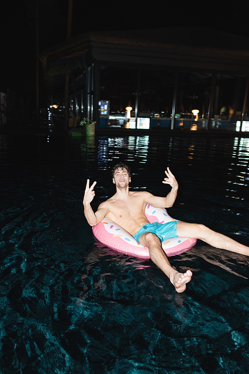 Selective focus of shirtless man showing peace sign on swim ring in pool at night