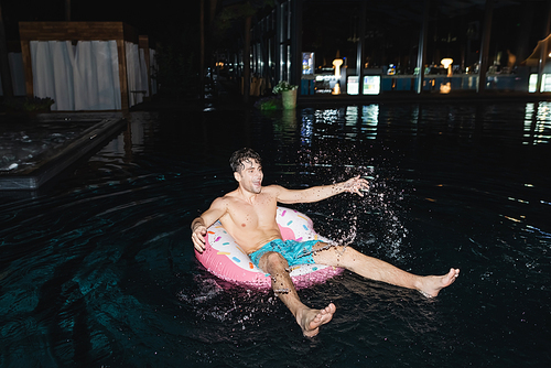 Selective focus of shirtless man on swim ring splashing water in pool at night