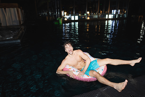 Excited muscular man sitting on swim ring in pool at night