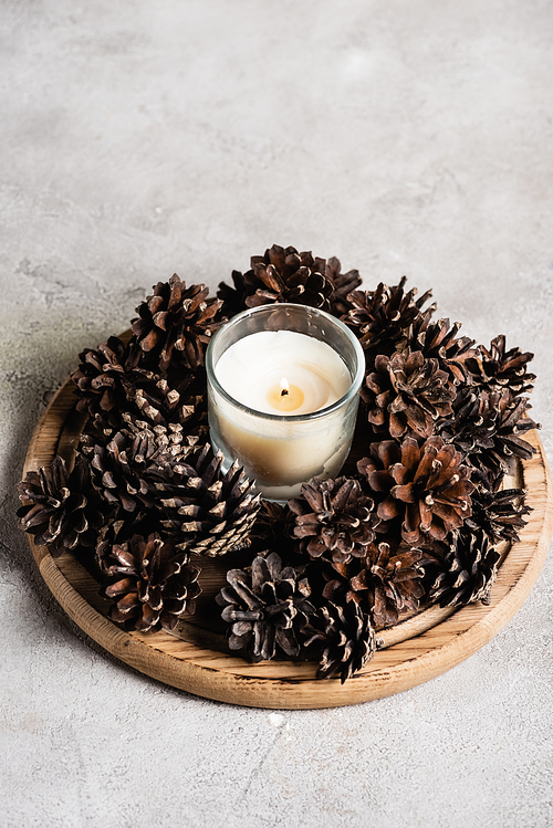 Scented candle with pine cones on wooden plate on grey and textured background