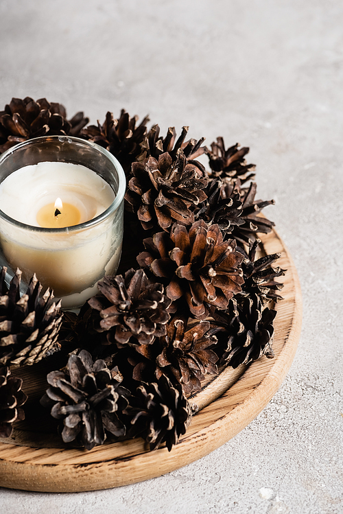 Scented candle with pine cones on wooden plate on textured and grey background