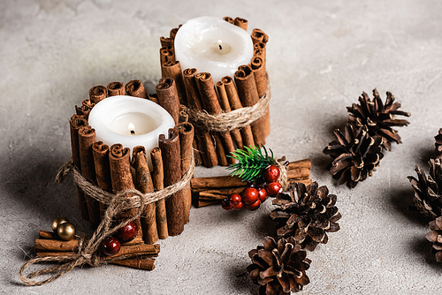 Decorated scented candles near pine cones on textured and grey background