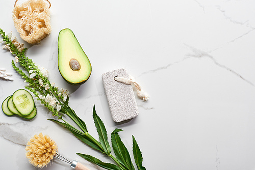 top view of branch with flowers, fresh avocado and cucumber near body brush, loofah and pumice stone on marble surface