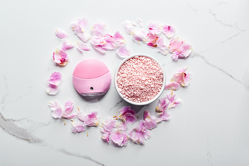 top view of silicone cleansing facial brush and bath salts on marble surface with pink petals