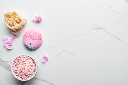 top view of silicone cleansing facial brush, bath salts and loofah on marble surface with pink petals