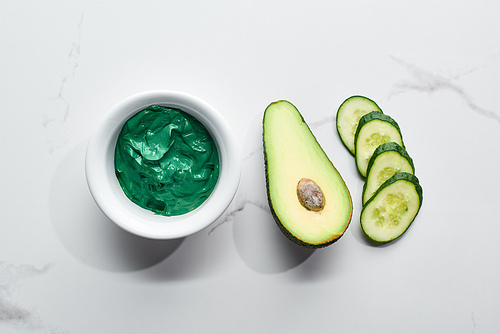 top view of natural clay mask near fresh avocado half and cucumber slices on marble background