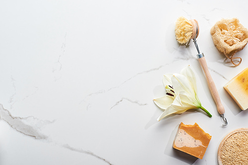 top view of natural soap pieces, body brush, loofah and face sponge near fresh flowers on marble surface