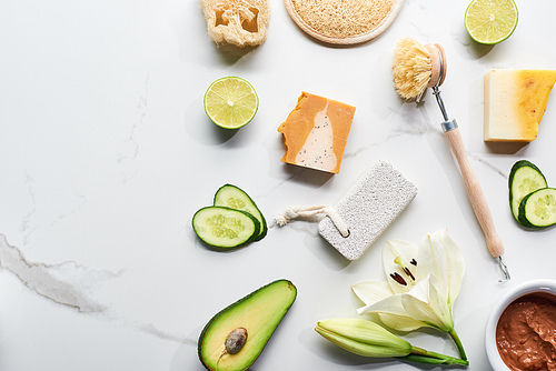 top view of natural soap pieces, body brush, loofah and pumice stone near fresh flower, lime, cucumber and avocado on marble surface