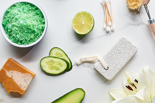 top view of green bath salts, soap and pumice stone near fresh flower, lime and cucumber on marble surface