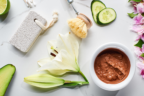 top view of body brush, pumice stone and natural clay mask near fresh flower, lime and cucumber on marble surface