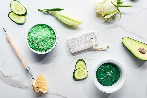 top view of body brush, pumice stone, bath salts and face mask near fresh avocado half, cucumber slices and flower on marble background