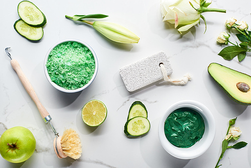 top view of natural clay mask, brush, pumice stone near apple, lime, cucumber, avocado and flowers on white surface