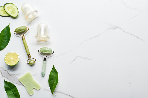 top view of nephrite massage rollers, vacuum jars, spatula, leaves, cucumber and lime on marble surface