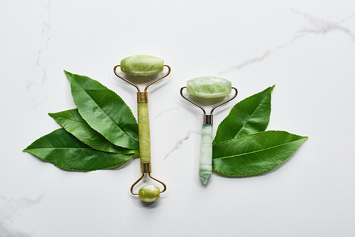 top view of green facial rollers and leaves on marble surface