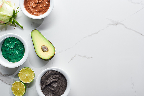 top view of cups with natural beauty products near flower, lime and avocado on marble surface