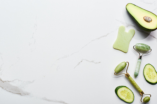 top view of massage rollers and pieces of cucumber and avocado on marble surface