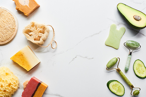 top view of facial rollers, loofah, bath sponges near cucumber and avocado on marble surface