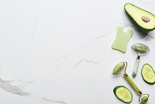 top view of facial spatula and rollers near avocado and cucumber on marble surface