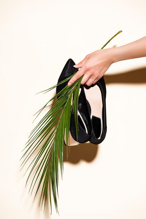 cropped view of woman holding black elegant shoes and palm leaf on beige background
