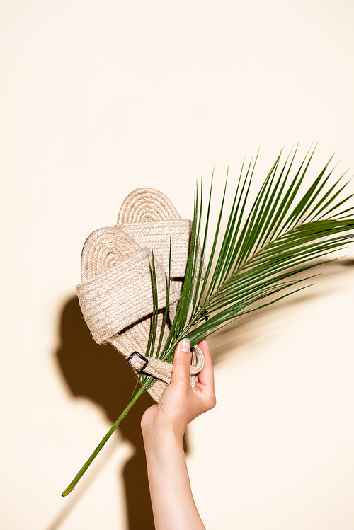 cropped view of woman holding summer braided sandals and palm leaf on beige background
